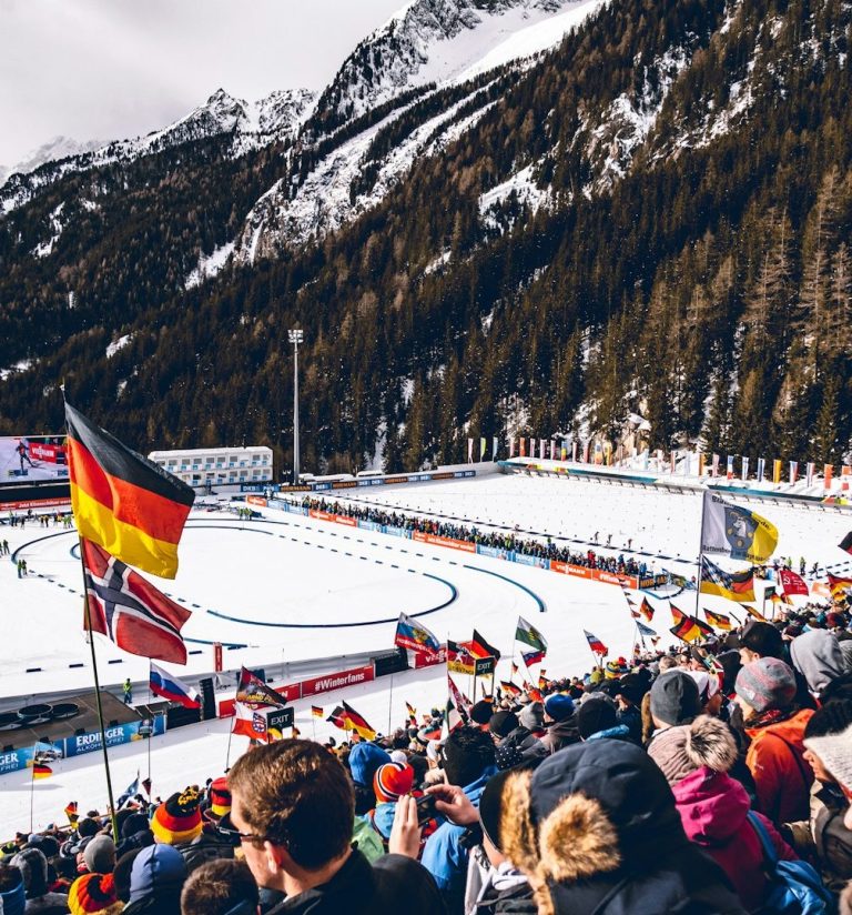 Menschenmenge in den Zuschauerrängen eines Biathlonevents mit Fahnen und schneebedeckter Landschaft.